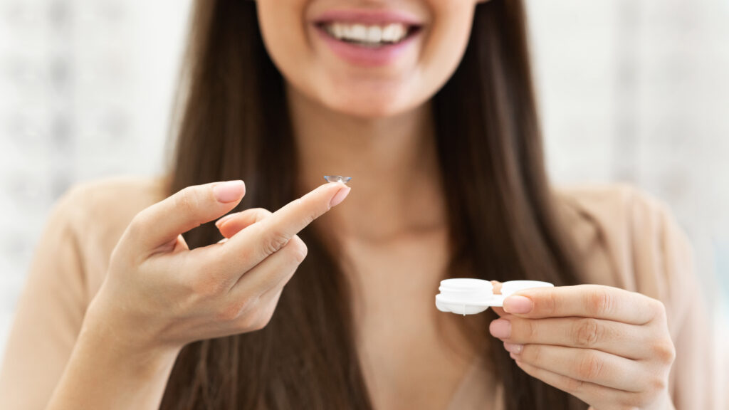 Closeup portrait of smiling woman showing contact eye lens to camera, holding it on finger, standing with plastic container case.